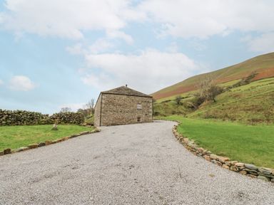 A gravel driveway leading to a stone building with grassy fields and hills around at Hollowgill Barn in Sedbergh