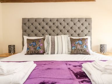 A bedroom with a grey tufted headboard white bed linens purple blanket decorative pillows and towels on the bed at Wheal Prosper@The West Dry in St Austell