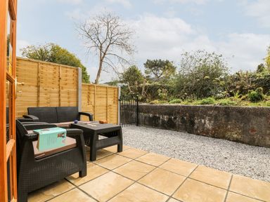 An outdoor patio with wicker furniture including a sofa, chair, and table surrounded by a wooden fence and garden wall at Wheal Prosper@The West Dry in St Austell
