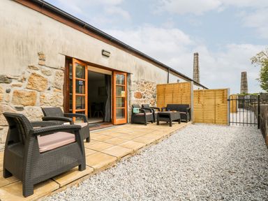 An outdoor patio area with black wicker chairs and a table on tiled flooring beside a stone and plaster wall at Wheal Prosper@The West Dry in St Austell