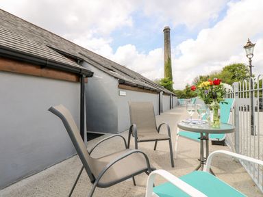 An outdoor seating area with chairs a small round table with wine glasses and flowers beside gray buildings with a chimney in the background at Wheal Prosper@The West Dry in St Austell
