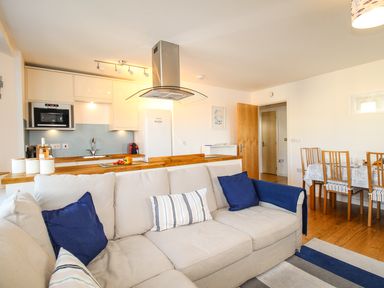 An open plan living area with a beige sofa with blue pillows a wooden dining table with chairs and a kitchen with white cabinets and appliances at Broadleys in Swanage