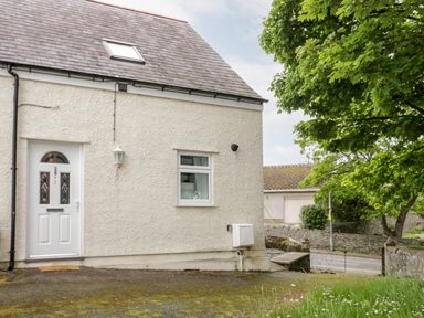 The exterior of a cream colored house with a white door and window near a road and a tree at 1 Old Kingdom Hall in Amlwch