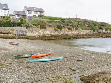 A rocky shoreline with four kayaks near a small body of water and houses on a hill at 1 Old Kingdom Hall in Amlwch