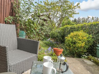 A garden with bushes and flowers a gray wicker chair and a glass table holding two white mugs and a French press at Red Roof in Marsh near Combe St Nicholas