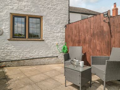 An outdoor patio area with two woven chairs a small glass-top table with a coffee press and mug a wooden fence and white textured wall at Red Roof Marsh near Combe St Nicholas