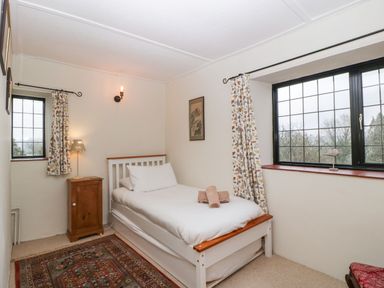 A bedroom with a single bed nightstand lamp two windows and a patterned rug at Red Roof Marsh near Combe St Nicholas