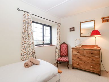 A bedroom with a bed, floral curtains, a wooden dresser with a mirror and lamp, and a patterned red chair at Red Roof in Marsh near Combe St Nicholas