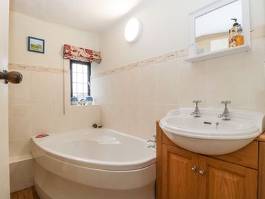A bathroom with a corner bathtub a sink with wooden cabinet and a small window with a floral blind at Red Roof Marsh near Combe St Nicholas