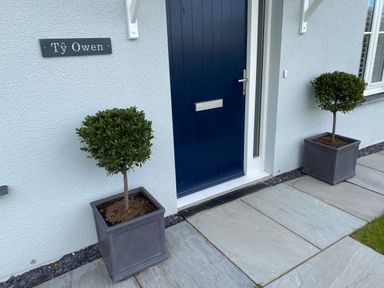 A blue front door with two potted topiary trees on either side and a sign reading Ty Owen on a white textured wall at Ty Owen Newport Pembrokeshire