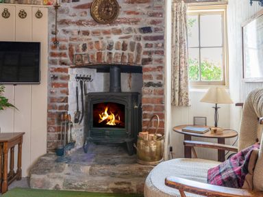A room with a lit wood stove set in a brick fireplace a wooden chair with a cushion a small table with a lamp and a window at End Cottage in Malborough