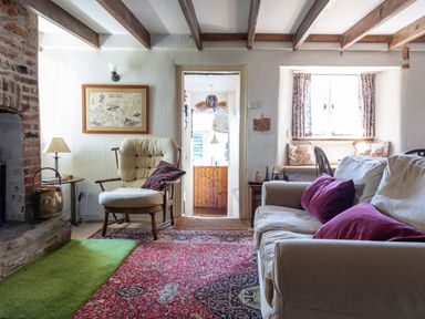 A living room with a beige sofa with purple cushions an armchair a red patterned rug and a window seat at End Cottage in Malborough