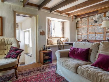 A living room with a beige sofa with red pillows a wooden chair a small wooden side table a window with curtains and a dining area at End Cottage in Malborough