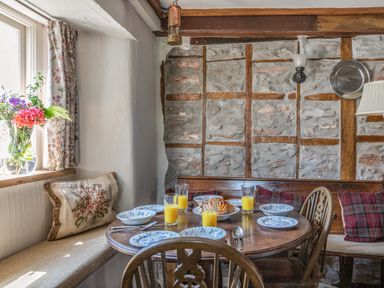 A dining area with a round wooden table set with plates and glasses of orange juice and a stone wall with wooden beams at End Cottage in Malborough