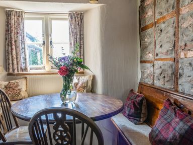 A small dining area with a round wooden table a vase with flowers wooden chairs a window with curtains and a bench with cushions at End Cottage in Malborough