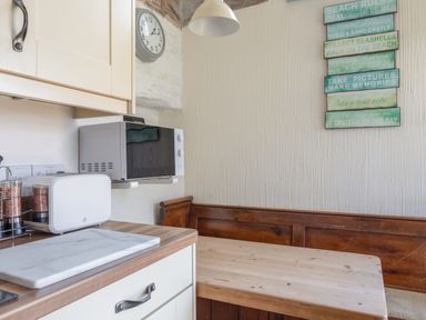 A kitchen corner with a wooden table and bench seating a microwave and toaster on the counter and a wall clock at End Cottage in Malborough