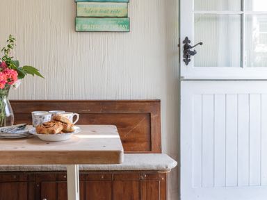 A wooden table with pastries on a plate cups and a vase of flowers next to a wooden bench and a white door at End Cottage in Malborough