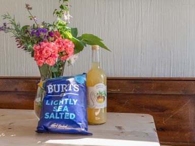 A table with a vase of flowers a bottle of juice and a packet of Burts lightly sea salted crisps at End Cottage in Malborough