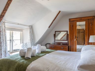 A bedroom with a bed covered in white sheets and a green blanket with rolled towels on it near a window with floral curtains and a wooden wardrobe with a mirror