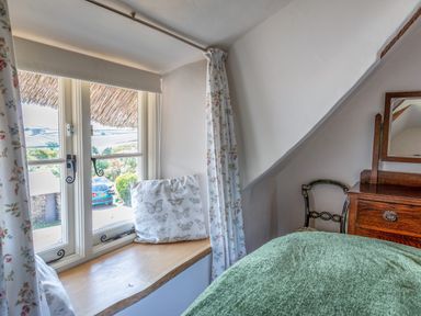 A bedroom corner with a window seat and cushions next to a wooden dresser and chair at End Cottage in Malborough