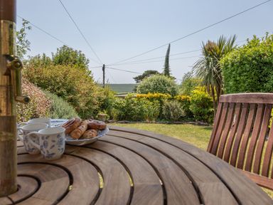 A wooden round table with cups and pastries in a garden with bushes and a lawn at End Cottage in Malborough