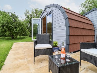 A patio with two wicker chairs and a small table with a bottle and glasses outside a small cabin at Meadow Lodge @ Berrys Place Farm in Churcham