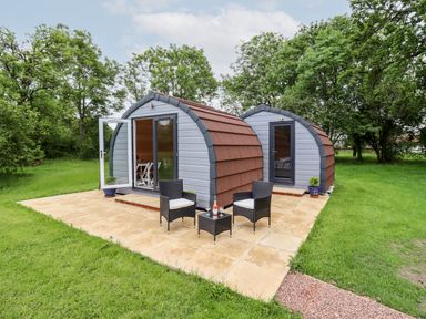 Two small curved lodges with doors and windows on a paved patio with two chairs and a table at Meadow Lodge @ Berrys Place Farm in Churcham