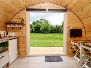 The interior of a wooden pod with kitchen appliances and a dining table facing an open door to a green lawn at Meadow Lodge @ Berrys Place Farm in Churcham