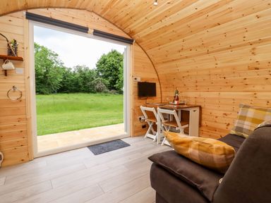 A wooden cabin interior with a small dining table and two chairs a couch with pillows and an open doorway showing a grassy outside area at Meadow Lodge @ Berrys Place Farm in Churcham