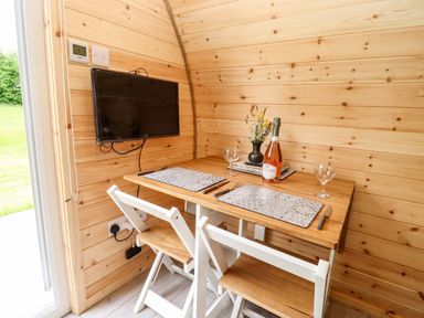 A small dining area with a wooden table set for two with placemats, wine glasses, a bottle of wine, and a vase with flowers next to a wall-mounted TV in a wooden room at Meadow Lodge @ Berrys Place Farm in Churcham