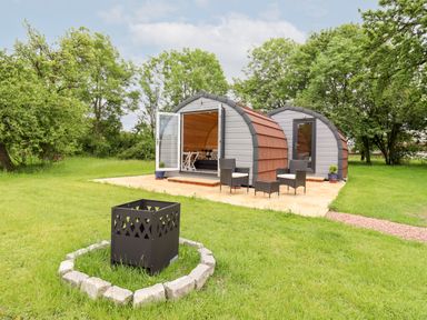 A pair of small curved lodges with outdoor chairs and a fire pit on a grassy lawn at Meadow Lodge @ Berrys Place Farm in Churcham