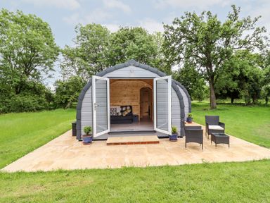 An outdoor cabin with open double doors showing a wooden interior and a sofa outside seating with chairs and a table on a paved patio surrounded by grass and trees at Meadow Lodge @ Berrys Place Farm in Churcham