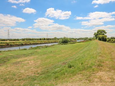 A grassy riverbank with a waterway and trees under a blue sky with clouds at Sweet Caroline Shepherd's Hut in Low Marnham near Sutton-On-Trent