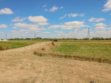 A grassy field with a dirt path under a blue sky with scattered clouds and electricity pylons in the distance at Sweet Caroline Shepherd's Hut in Low Marnham near Sutton-On-Trent