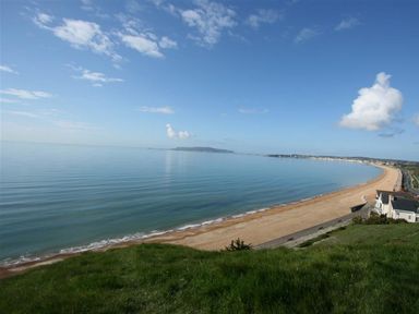 A beach with calm sea water and a clear sky viewed from a grassy hill at Mermaid House in Brewers Quay Harbour