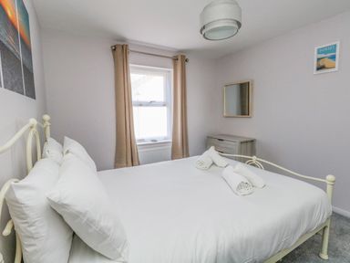 A bedroom with a white bedframe white bedding rolled towels a window with beige curtains a gray dresser and a wall mirror at Mermaid House Brewers Quay Harbour