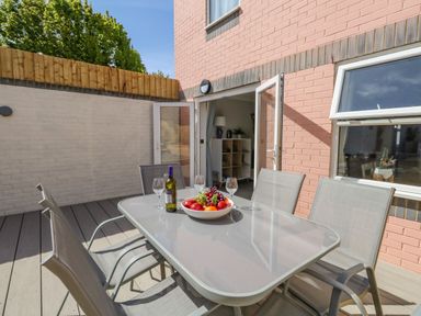 An outdoor patio with a glass table and six chairs a bowl of fruit a bottle of wine and glasses near an open door at Mermaid House in Brewers Quay Harbour