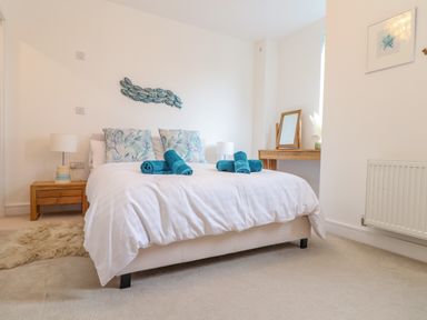 A bedroom with a double bed with white bedding and teal rolled towels a wooden bedside table with a lamp a wooden dressing table with a mirror and a framed picture on the wall at Upper Deck in Truro