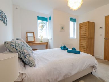 A bedroom with a double bed with blue pillows and rolled teal towels on it a wooden dressing table with mirror two windows with teal blinds and a wooden wardrobe at Upper Deck in Truro
