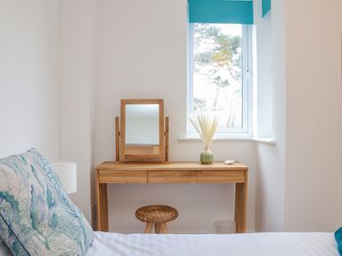 A bedroom corner with a wooden dressing table mirror a wicker stool a window with a blue blind and part of a bed with a blue patterned pillow at Upper Deck in Truro