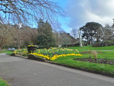 A paved path beside flower beds and trees in a park with sculptures at Upper Deck in Truro
