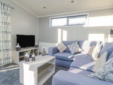 A living room with a blue sectional sofa a coffee table with vases a television on a stand and patterned curtains at Yealand 36 in Tewitfield