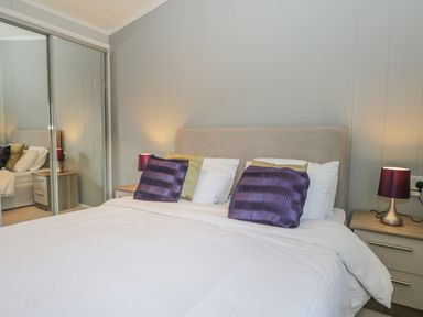 A bedroom with a bed with white bedding purple and green pillows two bedside tables with lamps and mirrored wardrobe doors at Yealand 36 in Tewitfield