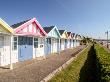 A row of colorful beach huts with pastel roofs and blue and white doors along a paved walkway near the sea at Portside Greenhill Weymouth