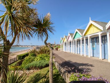 A row of colorful beach huts along a path with plants and a beach in the background at Portside Greenhill Weymouth