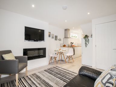A living room with a striped rug a wall-mounted TV a small dining table with two white chairs and a kitchen area at Portside Greenhill Weymouth