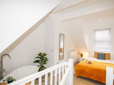 A bedroom with a bed, pillows, a blanket, a window, a nightstand with a lamp and plant, a full-length mirror, and a bathtub with plants nearby at Portside Greenhill Weymouth