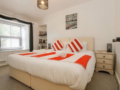 A bedroom with a large bed with red and white cushions and runner a nightstand and shelves with books at Walter in Newlyn
