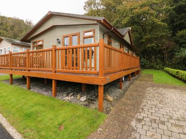 A wooden cabin with a raised deck and railing next to a paved driveway and grass at 16 Heritage Park in Heritage Park near Kilgetty
