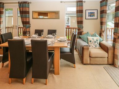A dining area with a wooden table set for six and an armchair with cushions near windows at 16 Heritage Park in Heritage Park near Kilgetty